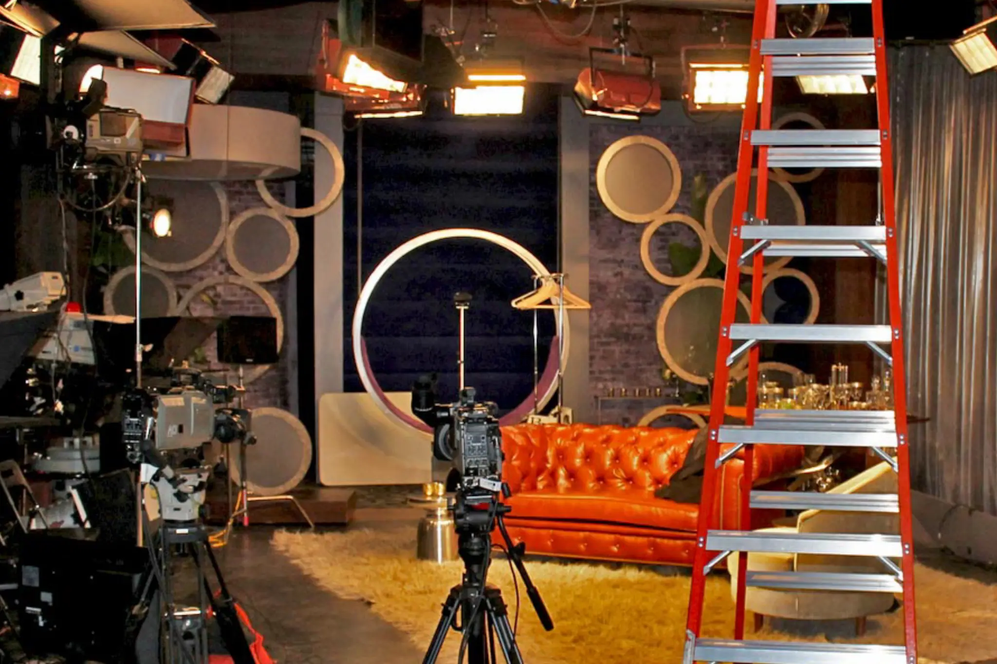 A television studio setup featuring a red ladder, cameras, an orange couch, and decorative wall circles under bright stage lights